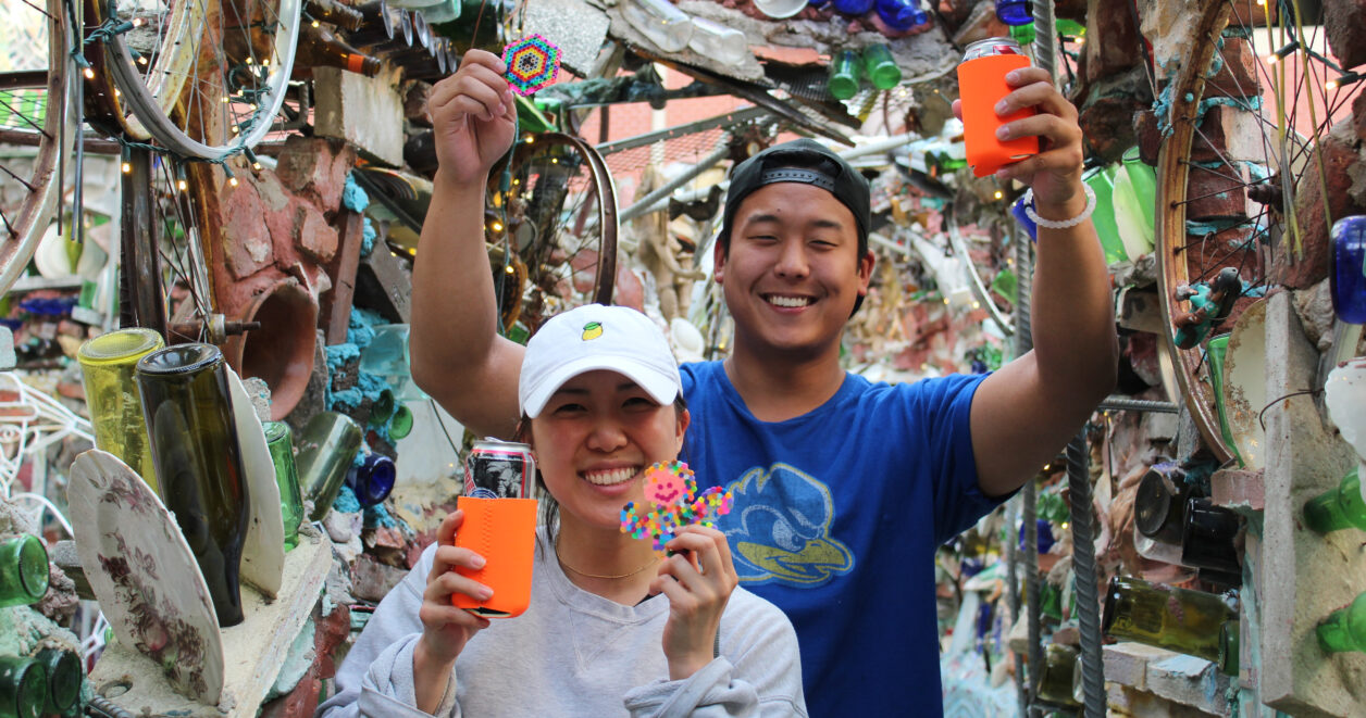 Two smiling people hold up a beer in a koozie in one hand and a perler bead creation in the other as they stand in a mosaicked hallway at Philadelphia's Magic Gardens.