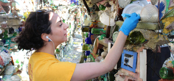 A woman wearing a yellow t-shirt and blue gloves uses a small brush to clean between two plates on a wall at Philadelphia's Magic Gardens