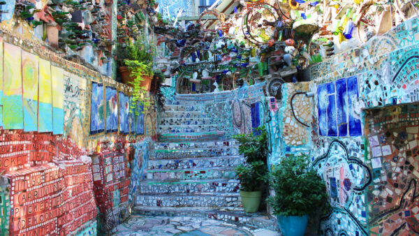 Outside at Philadelphia's Magic Gardens a staircase goes up between two flat walls. The stairs and walls are covered in colorful tiles. Above the staircase and flat walls the walls continue but are made out of objects such as bike wheels and bottles. 