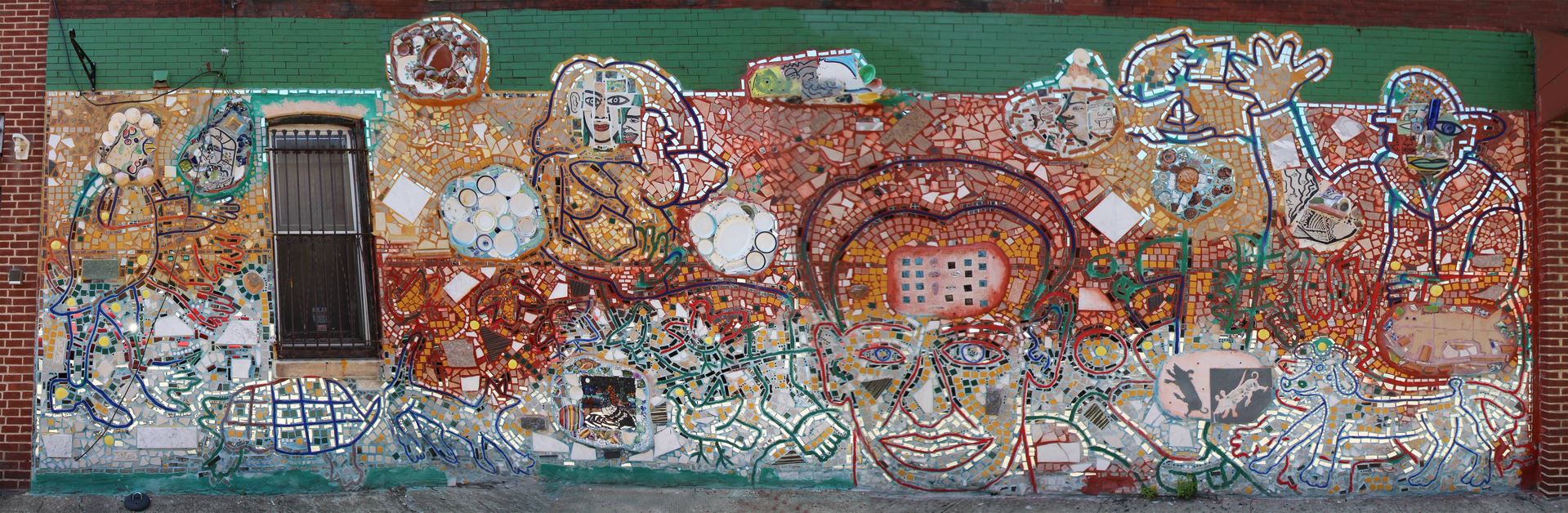A horizontal image of a colorful mosaic mural by Isaiah Zagar. It features the outlines of people and animals, and uses primarily red and yellow grout.