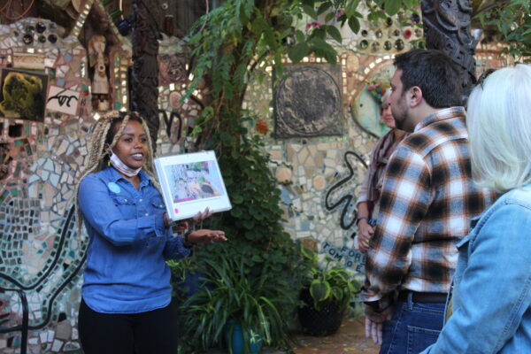 A tour guide smiles and is showing a photograph to a group of tour-takers outside at Philadelphia's Magic Gardens. Plants and a mosaicked wall can be seen in the background. 