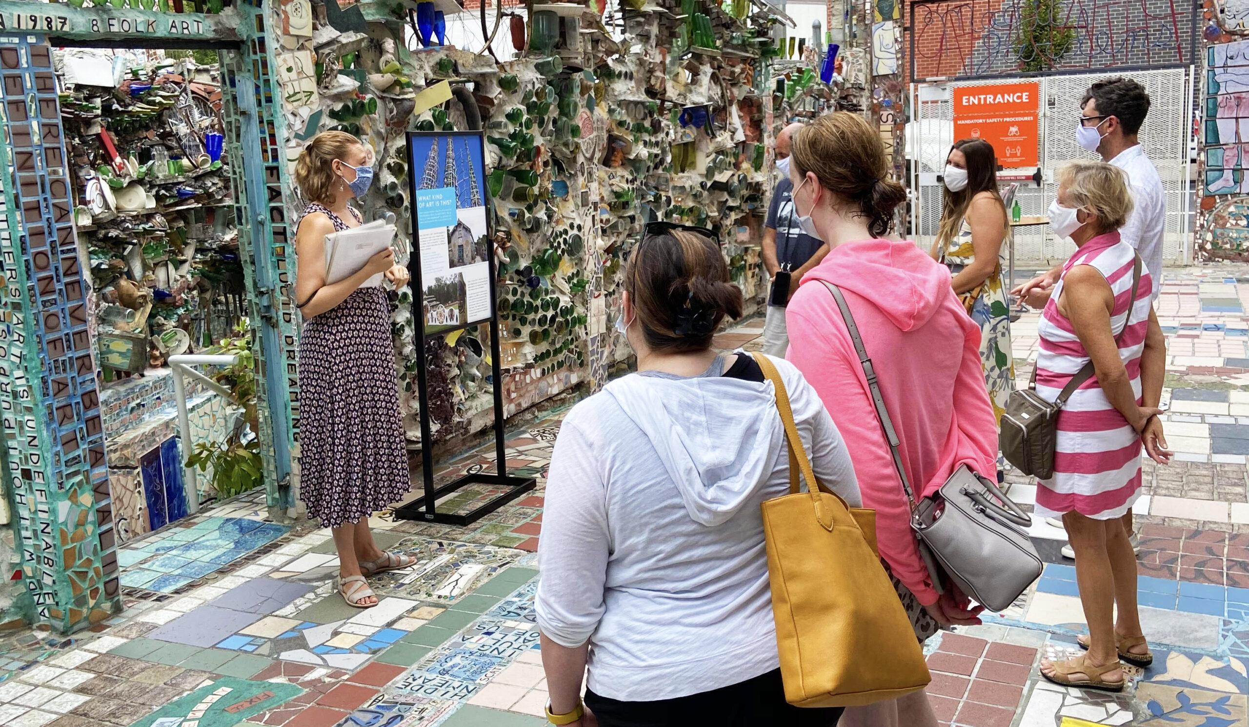 A tour guide wearing a dress and mask stands in front of a small group of people in the mosaicked courtyard of Philadelphia's Magic Gardens. She holds a stack of visual aids.