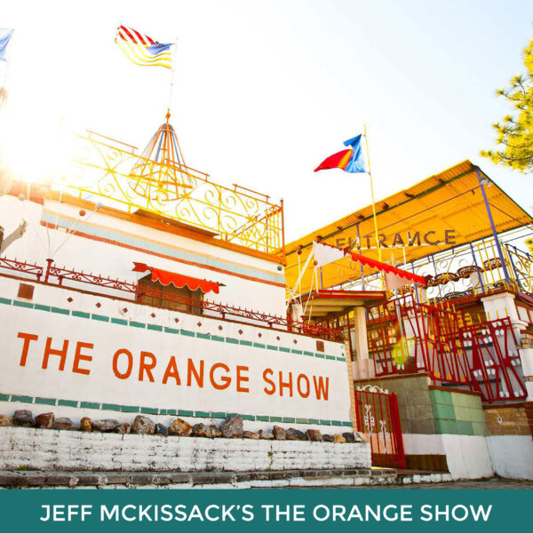 Jeff McKissack’s The Orange Show. A large white building with a yellow awning outside. The words "The Orange Show" are painted in orange along the side of the building. 