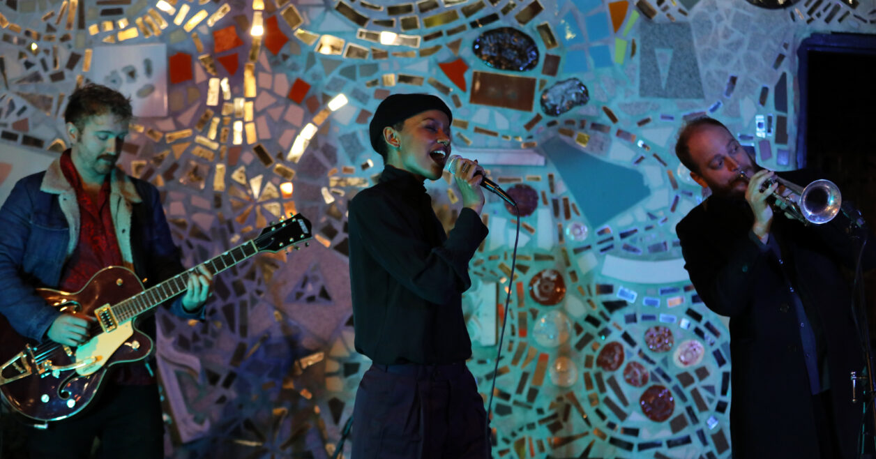 Against a backdrop of mosaics a singer, guitarist, and trumpeter perform.