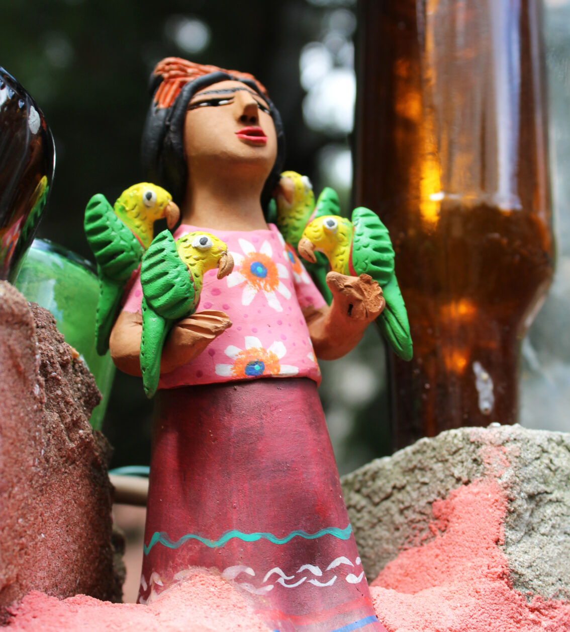 A ceramic folk art statuette depicting a woman in a pink dress holding yellow and green parrots.