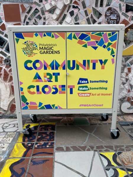 The Community Art Closet, a low, rolling cabinet, in front of mosaics. Text on the cabinet reads "Take Something, Make Something, Create Art at Home!"
