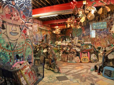 A look into the Watkins Street Studio. It has high ceilings with red steel beams and chandeliers created from found objects. On the ground are low walls made of found objects, and the floor is tiled mosaic.