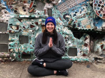 A young woman sits cross-legged with prayer hands in front of a mosaicked wall.