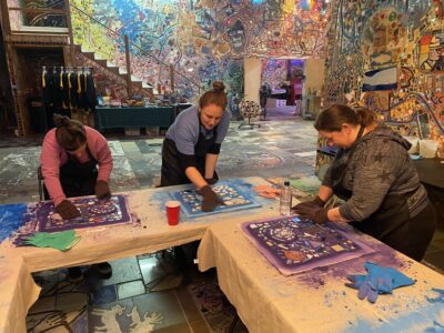 Three women wearing aprons and gloves lean over tables working on colorful mosaics.