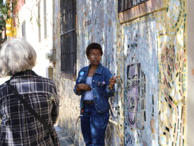 A woman tour guide gestures to a mosaic mural behind her.