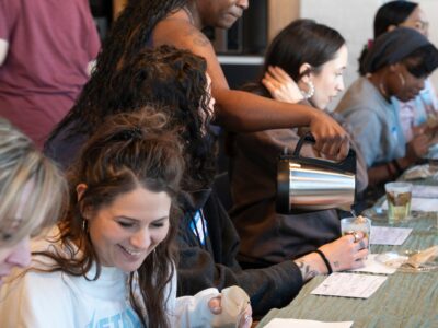 A woman pours tea into the mug while a group of people sit around a long table smiling.