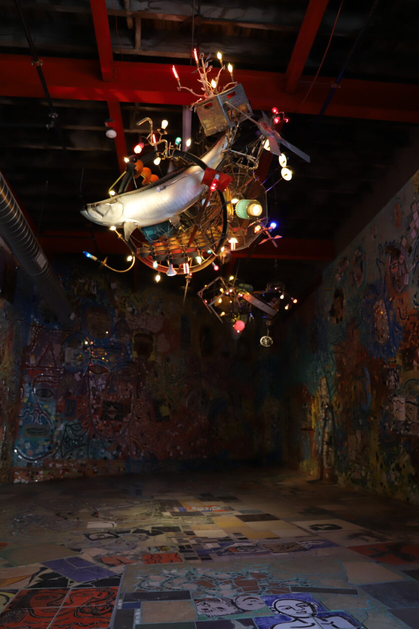 The Magic Gardens Studio. A high-ceilinged room with mosaicked walls and floor. A large chandelier made from found objects and a taxidermy fish hangs in the center.