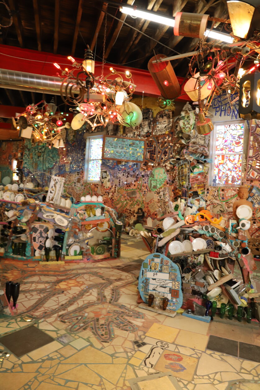 The Magic Gardens Studio. A high-ceilinged room with mosaics on the walls and floor. Low sculptural walls made of bottles and plates are in the center.