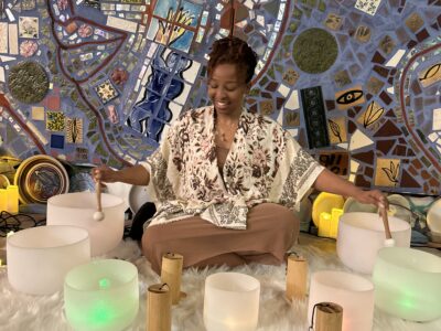 A woman sits cross-legged on a blanket and plays glass bowls in front of a mosaicked wall.