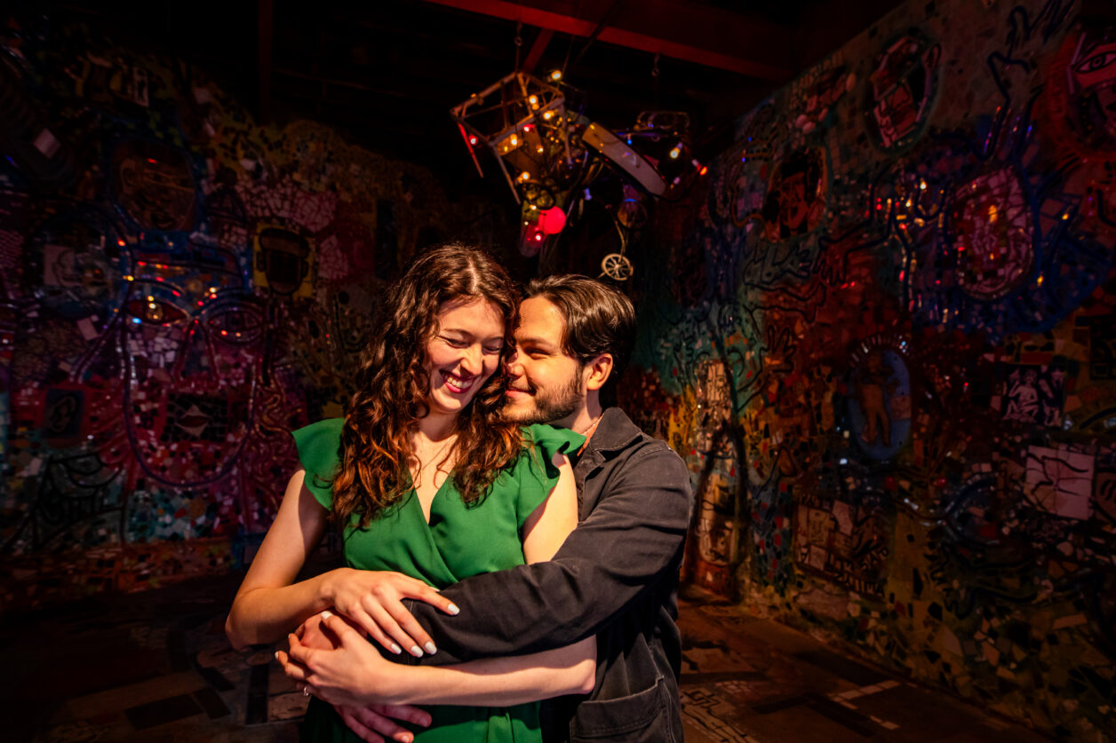 A couple hugs and smiles in the middle of a room with mosaicked walls.