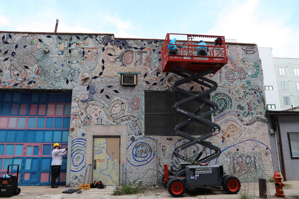 Three PMG staff members work on removing tiles from the façade of a mosaicked building using a forklift and chisels.
