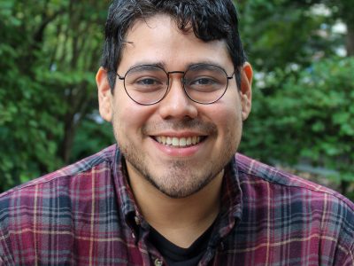Artist Santiago Galeas smiles in a plaid shirt with trees in the background.