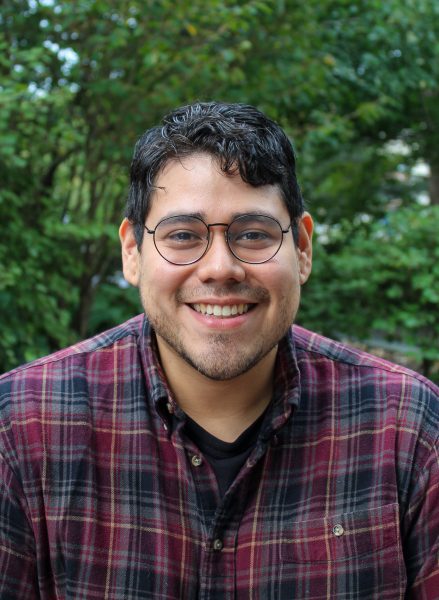 Artist Santiago Galeas smiles in a plaid shirt with trees in the background.