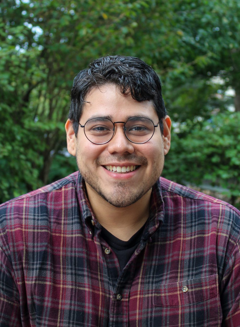 Artist Santiago Galeas smiles in a plaid shirt with trees in the background.