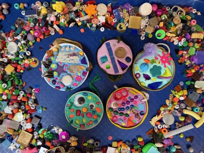 Five jar lids on a dark blue background are filled with brightly colored grout. The grout is embedded with beads, googly eyes, neon colored fish gravel, seashells, tiles, glass, sequins, and other colorful things. The artworks are surrounded by a pile of loose materials like those used in the grout.
