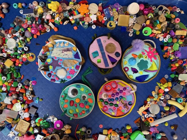 Five jar lids on a dark blue background are filled with brightly colored grout. The grout is embedded with beads, googly eyes, neon colored fish gravel, seashells, tiles, glass, sequins, and other colorful things. The artworks are surrounded by a pile of loose materials like those used in the grout.