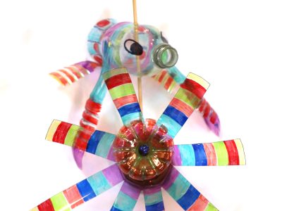Two brightly colored garden ornaments made out of plastic bottles. One look like a flying fish with a propeller on its head. The other looks like a pinwheel.